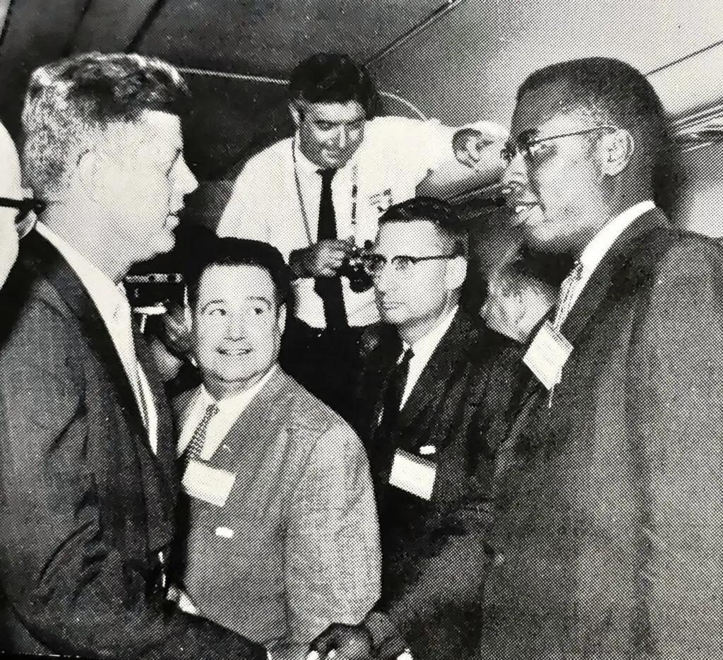 John F. Kennedy and Nathaniel S. Colley shake hands surrounded by people.