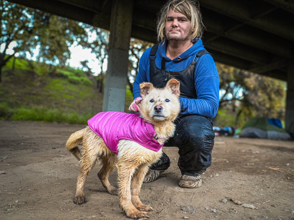 A homeless individual next to a dog in a pink jacket