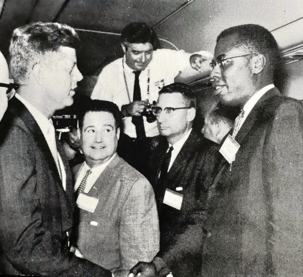 John F. Kennedy and Nathaniel S. Colley shake hands surrounded by people.