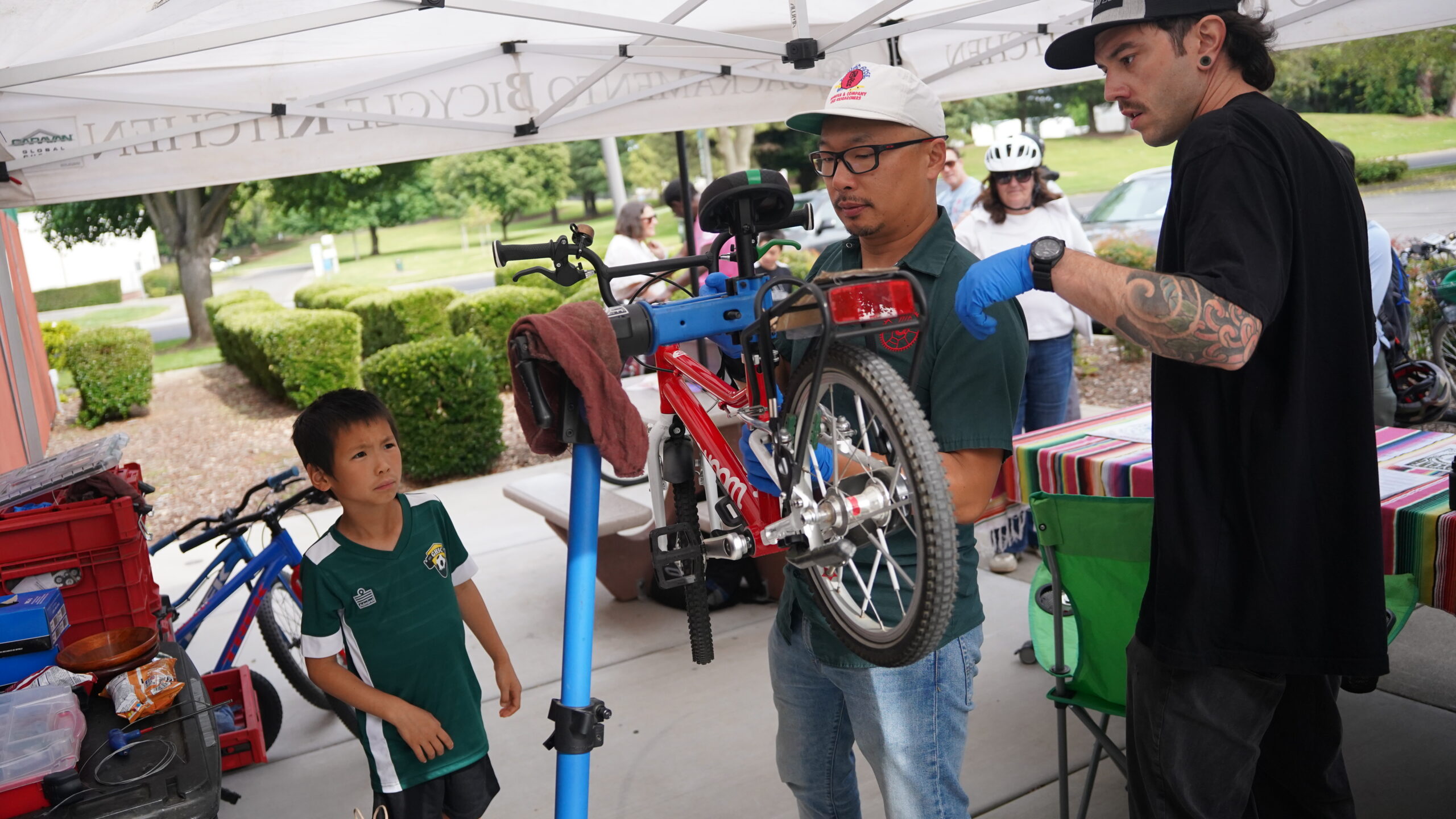A young boy watches as mechanics check the brakes on his red bicycle.