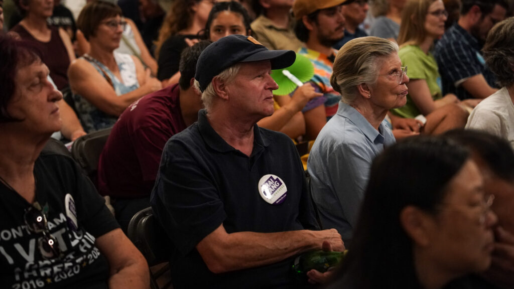 A person with short white hair wearing a black shirt and ball cap sits in a crowd listening.