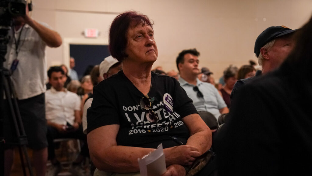 A woman with maroon hair is seated in a crowd, wearing a black shirt, and gazing off to the right.