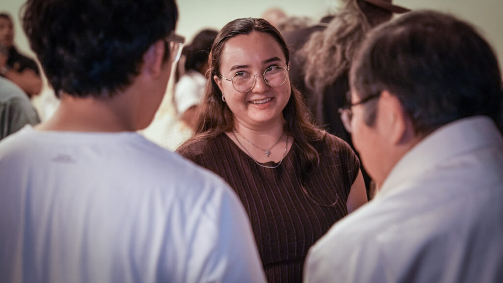 A person with long brown hair and wearing a brown shirt and glasses smiles while chatting with two people in light-colored shirts with short dark hair.