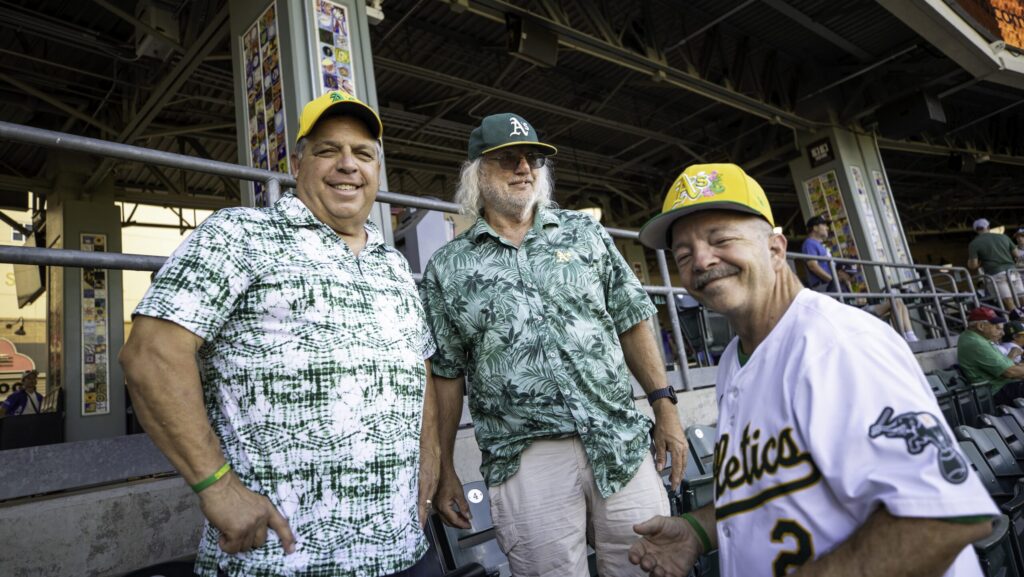 Three men wearing A's baseball hats stand next to each other.